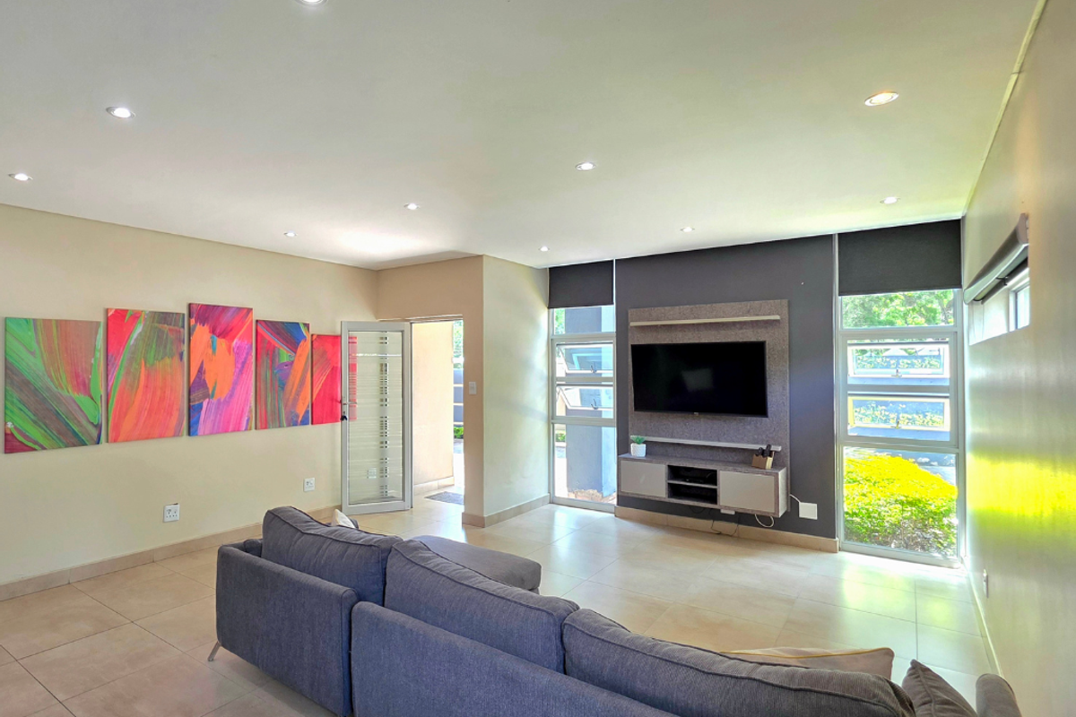 A bright and modern living room featuring tiled floors, recessed downlights, and a built-in media console with large windows overlooking the garden.