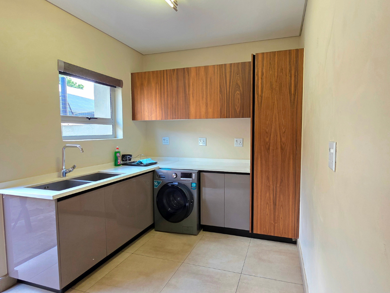 A clean and functional laundry room featuring modern built-in cabinetry, a stainless steel sink, and tiled flooring with space for a washing machine.