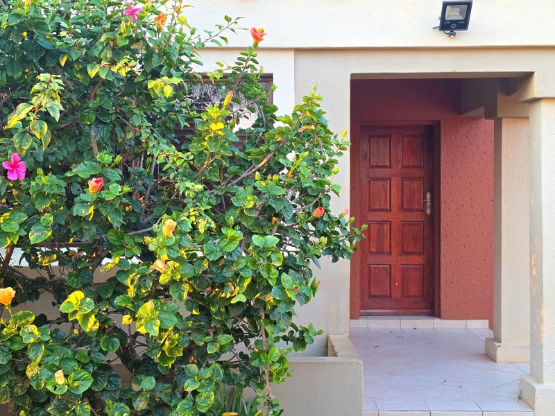 A welcoming front entrance featuring a solid wood panel door, tiled porch area, and exterior security lighting, framed by a vibrant flowering shrub.