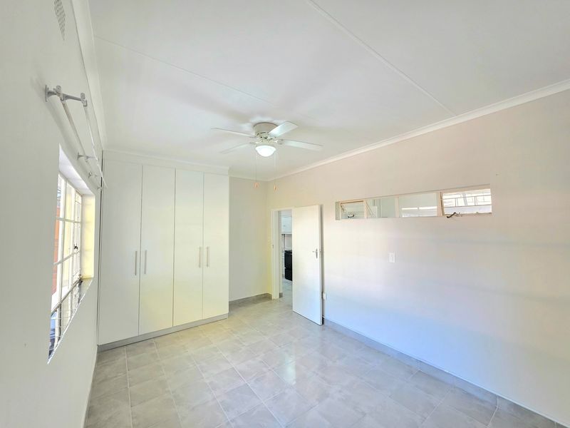 A bright, empty bedroom featuring neutral tiled flooring, built-in white cupboards, a ceiling fan, and natural light from steel-framed windows.