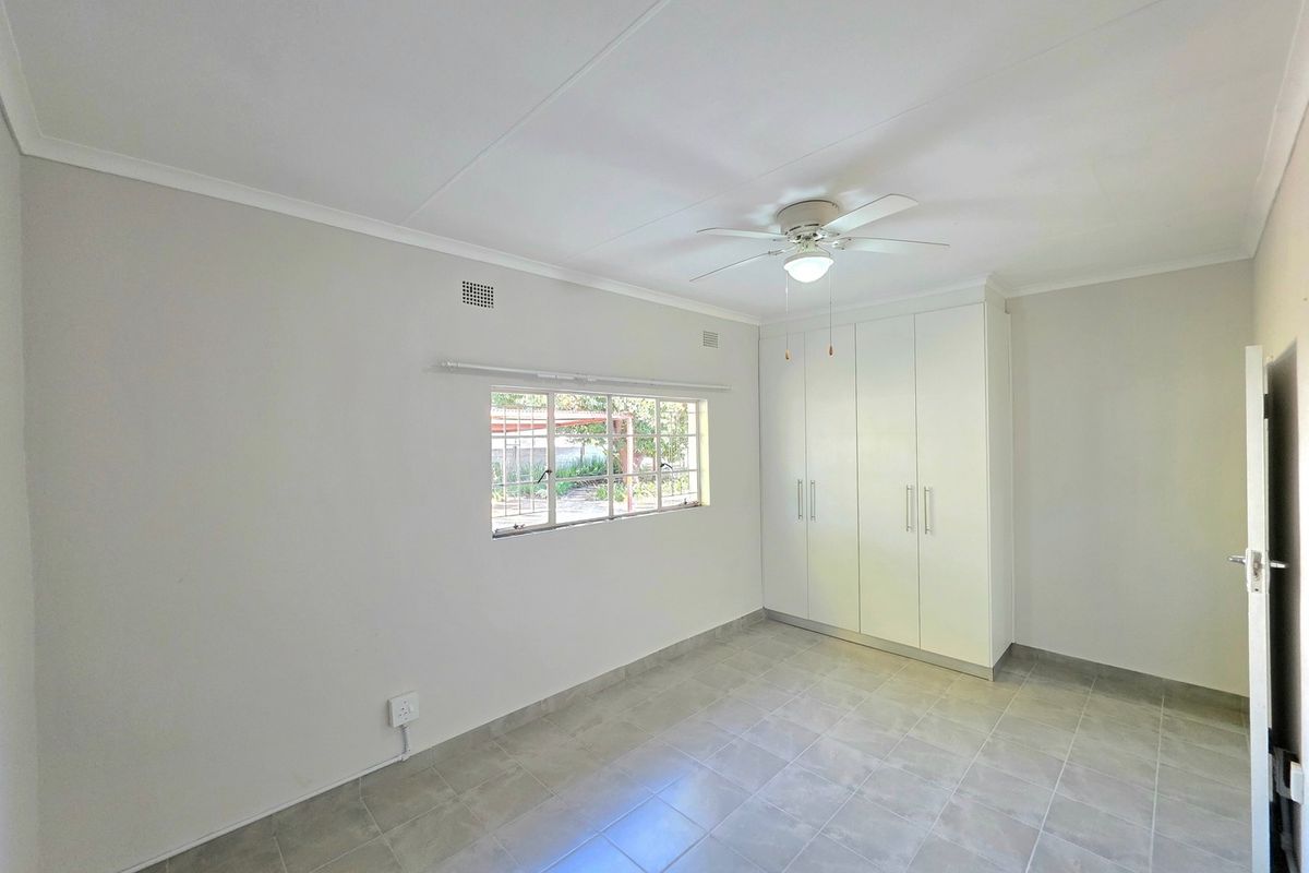 A bright, empty bedroom featuring neutral tiled flooring, built-in white cupboards, and a ceiling fan, offering a clean and versatile living space.