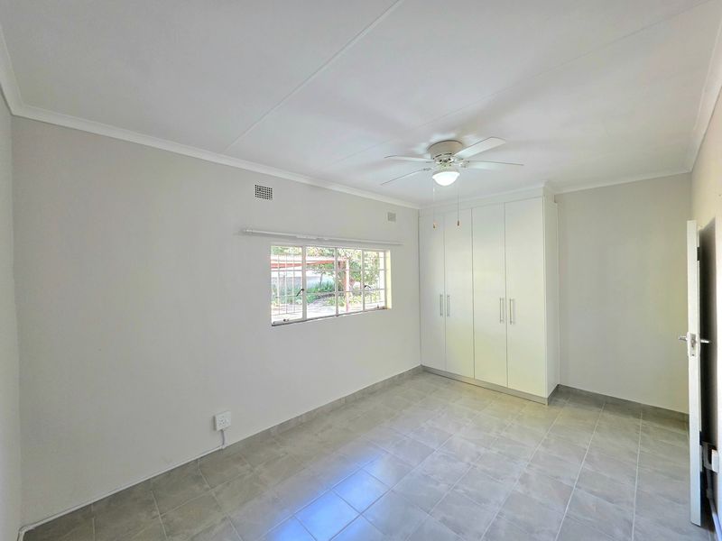 A bright, empty bedroom featuring neutral tiled flooring, built-in white cupboards, and a ceiling fan, offering a clean and versatile living space.