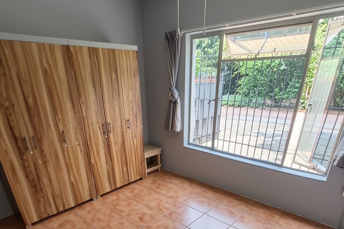 A bedroom featuring built-in wooden cupboards, tiled flooring, and a large window with security bars overlooking the exterior.