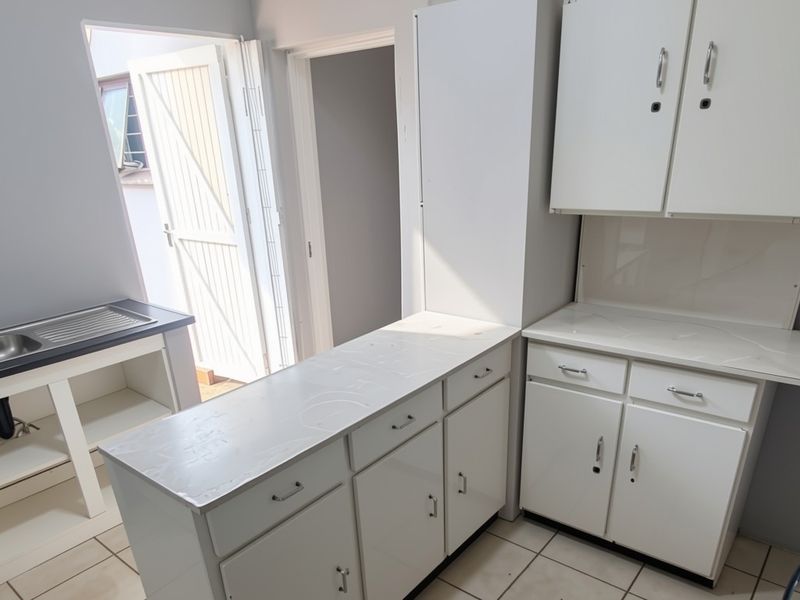 A functional kitchen area featuring white built-in cabinetry, laminate countertops, a stainless steel sink, and tiled flooring with direct access to an outdoor area.