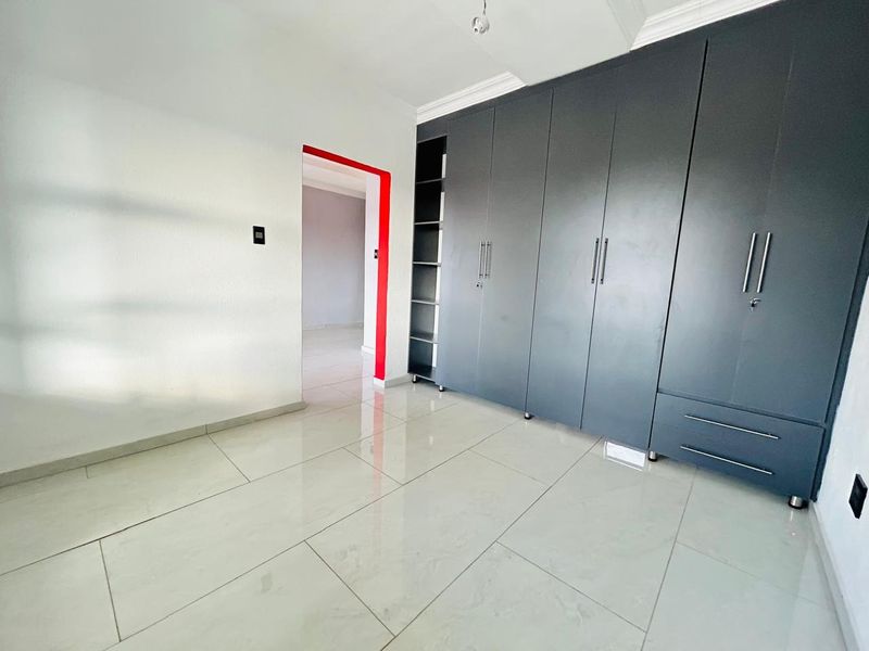 A bright, empty bedroom featuring modern grey built-in cupboards, polished tiled flooring, and clean white walls with a decorative red door frame accent.