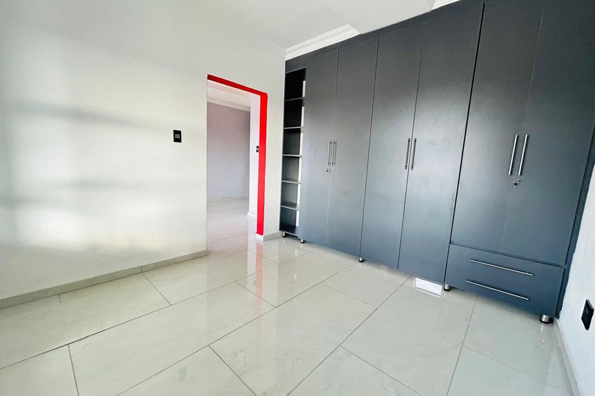 A bright, empty bedroom featuring modern grey built-in cupboards, polished tiled flooring, and clean white walls with a decorative red door frame accent.