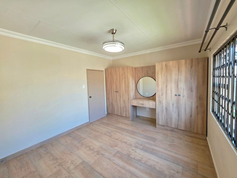 A bright, empty bedroom featuring modern wood-look laminate flooring, built-in cupboards with an integrated vanity, and a ceiling fan.