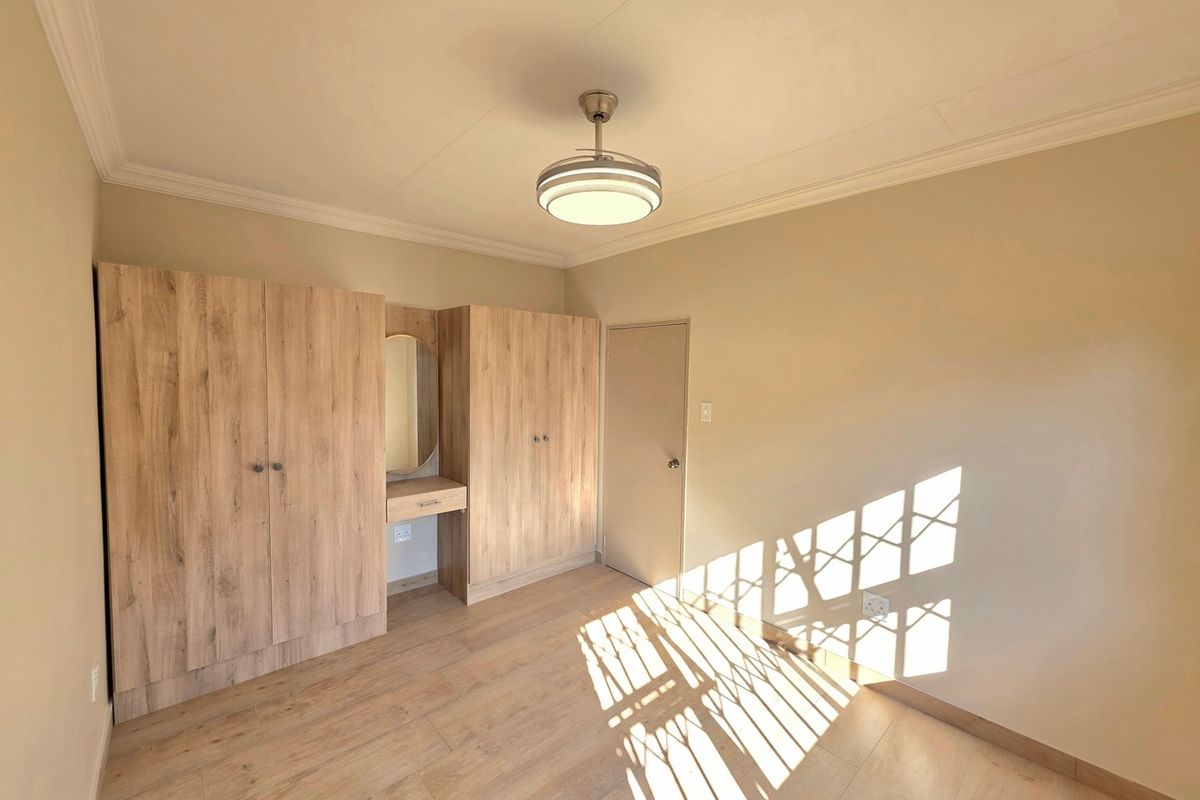 A bright, empty bedroom featuring light wood-look laminate flooring, built-in cupboards with an integrated vanity, and a modern ceiling light fixture.
