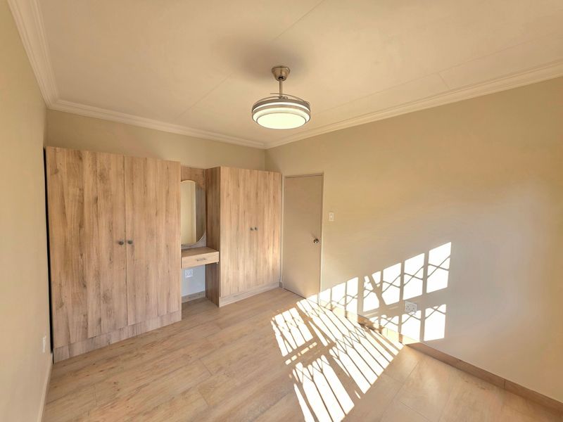 A bright, empty bedroom featuring light wood-look laminate flooring, built-in cupboards with an integrated vanity, and a modern ceiling light fixture.