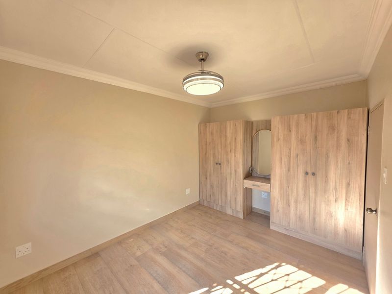 A bright, empty bedroom featuring light wood-look laminate flooring, neutral walls, and integrated built-in cupboards with a central vanity mirror.