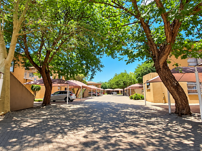 A well-maintained apartment complex exterior featuring a paved driveway, mature trees, and shaded parking bays for residents.