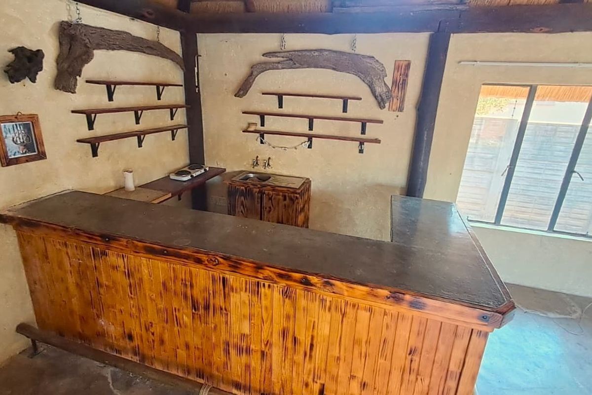Rustic bar area featuring a built-in wooden counter, stainless steel sink, and wall-mounted shelving, set against textured plaster walls with exposed timber beams.
