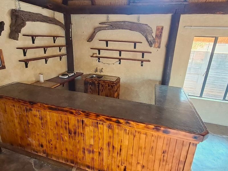 Rustic bar area featuring a built-in wooden counter, stainless steel sink, and wall-mounted shelving, set against textured plaster walls with exposed timber beams.