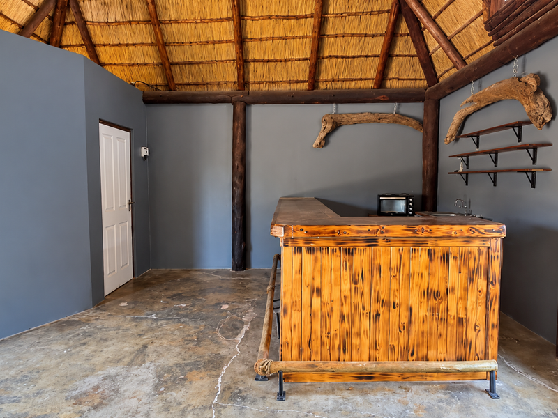 A rustic bar area featuring a thatched roof, exposed timber support beams, a built-in wooden bar counter with a sink, and concrete flooring.