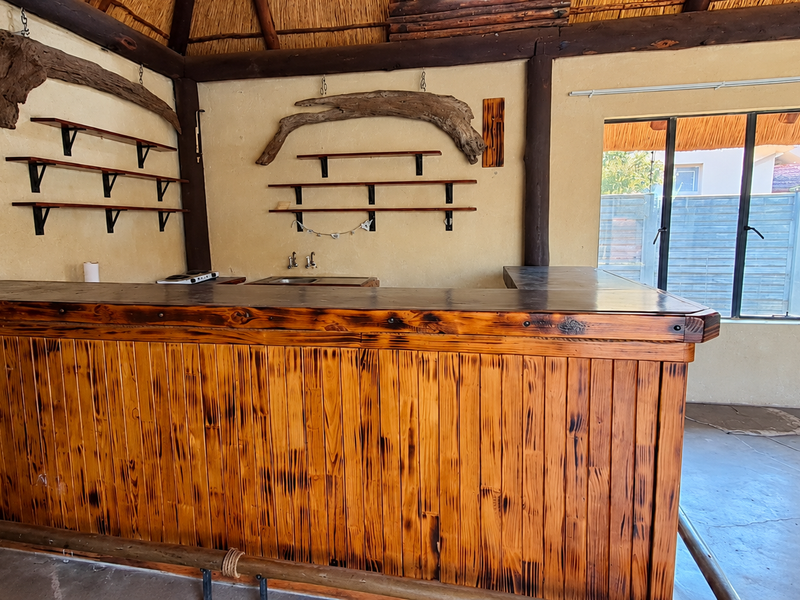 A rustic bar area featuring a built-in wooden counter, integrated sink, wall-mounted shelving, and exposed timber beams under a traditional thatched roof.