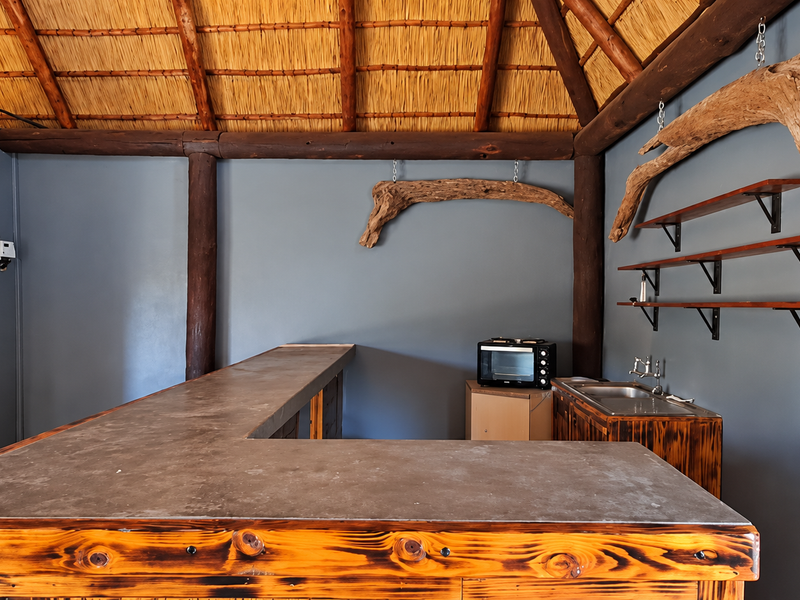 A rustic bar area featuring a thatched roof, exposed timber beams, a built-in bar counter, and a stainless steel sink with wall-mounted shelving.