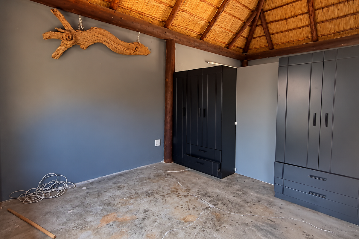 A bedroom featuring a traditional thatched roof, exposed timber support poles, and dark built-in wardrobes, set against a grey feature wall.