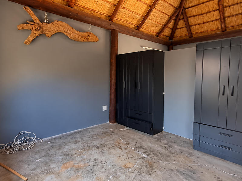 A bedroom featuring a traditional thatched roof, exposed timber support poles, and dark built-in wardrobes, set against a grey feature wall.