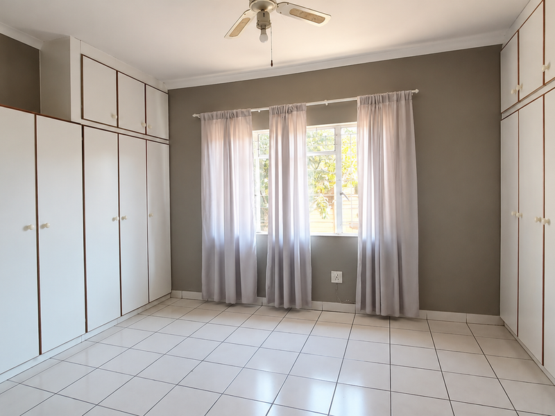 A bright, empty bedroom featuring extensive built-in cupboard storage, tiled flooring, and a ceiling fan, offering a clean and neutral space.