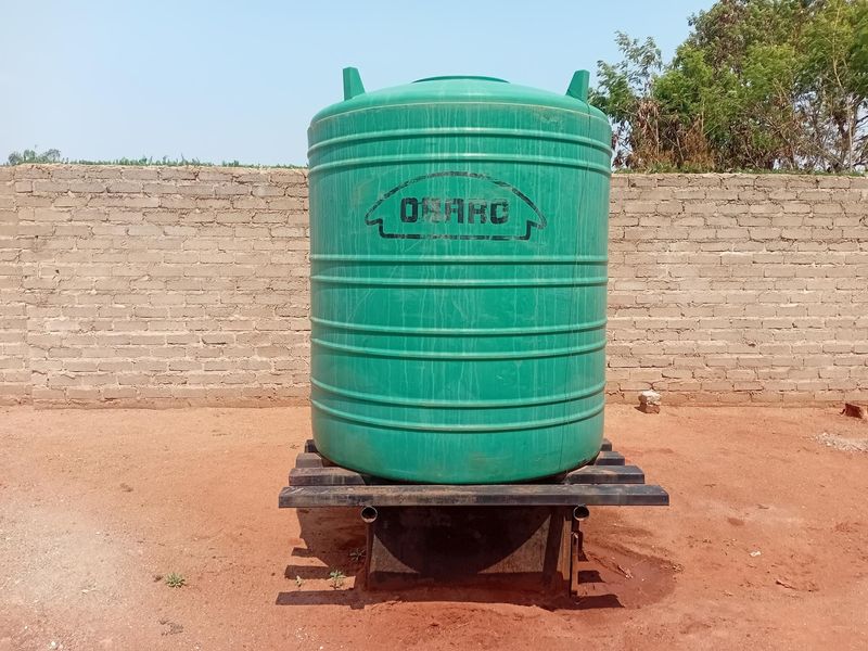 A green plastic water storage tank mounted on a metal support stand, situated in an outdoor area with a brick boundary wall.