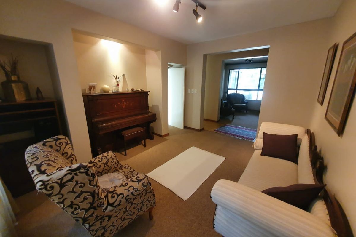 A living room area featuring recessed wall shelving, carpeted flooring, and track lighting, leading into an adjacent room with large windows.