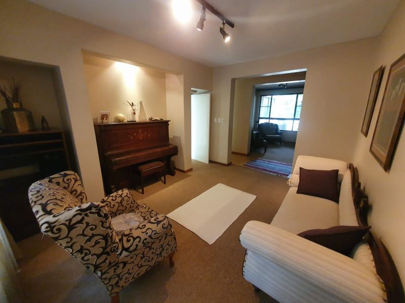 A living room area featuring recessed wall shelving, carpeted flooring, and track lighting, leading into an adjacent room with large windows.
