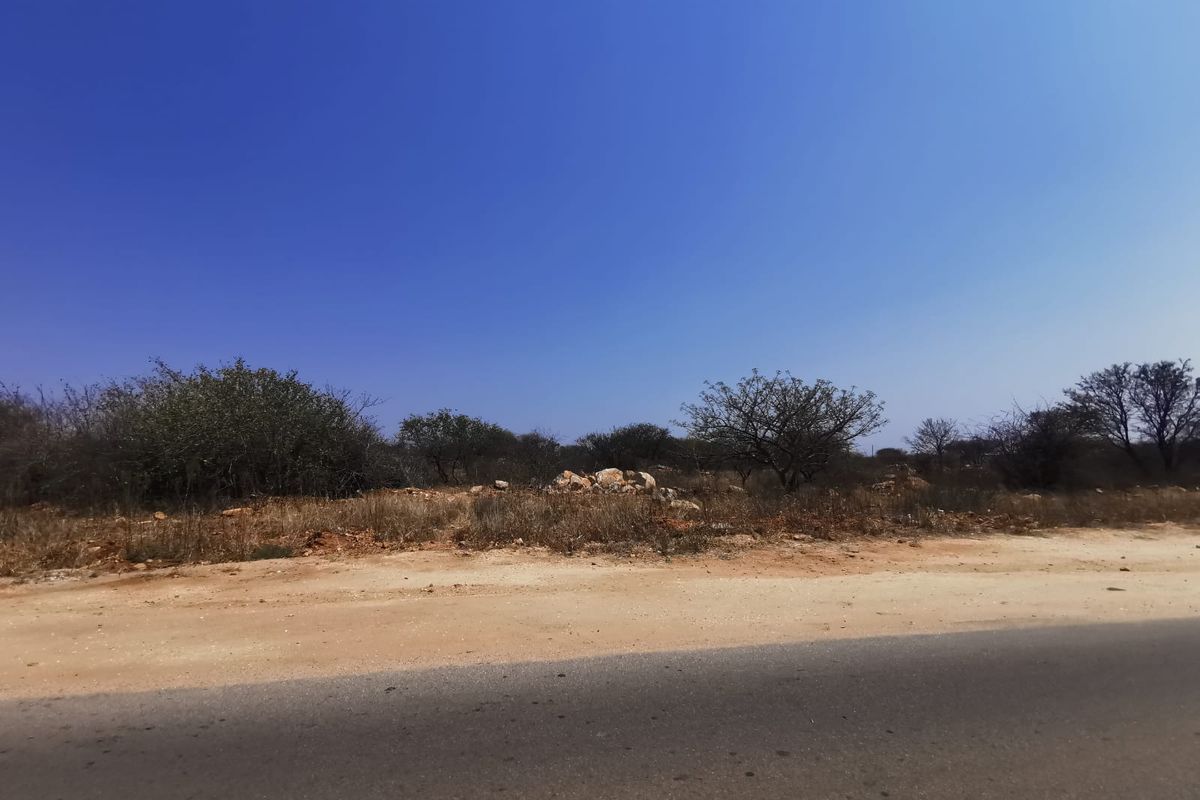 A view of a vacant residential plot from the adjacent paved road, featuring natural vegetation and rocky terrain.