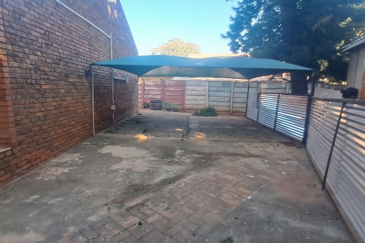 Rear exterior area featuring a shade sail, brick and concrete paving, and corrugated metal fencing, showing signs of wear and weathering.
