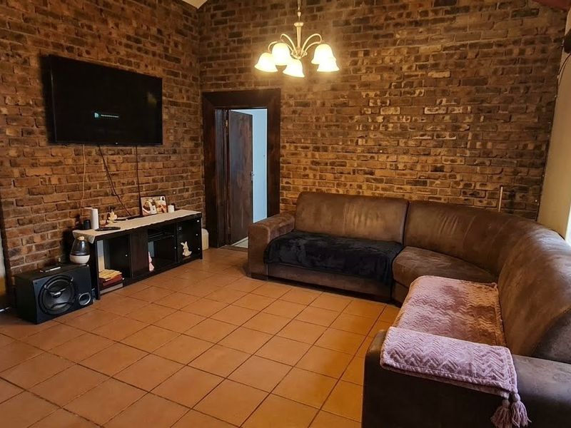 A living room featuring exposed brick walls, tiled flooring, and a vaulted ceiling, providing a rustic aesthetic with natural light from a large window.
