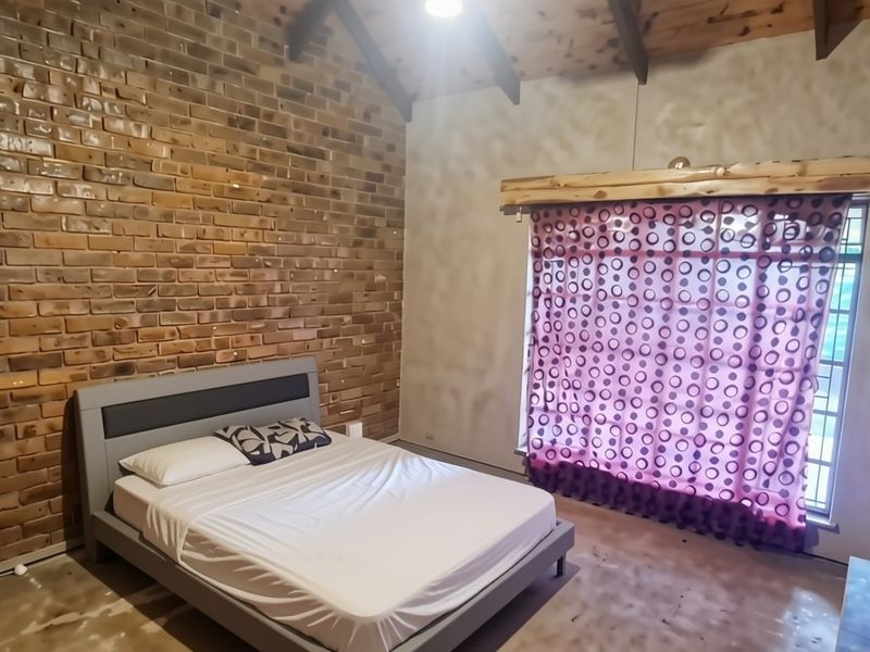A bedroom featuring an exposed brick feature wall, vaulted timber ceiling with exposed beams, and a large window providing natural light.