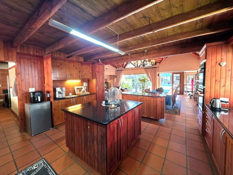 Spacious open-plan kitchen featuring timber cabinetry, a central island with stone countertop, exposed wooden ceiling beams, and terracotta floor tiles.
