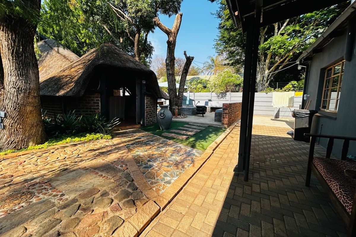 A spacious outdoor entertainment area featuring a thatched lapa, paved walkways, and a brick-walled patio space set within a mature garden.