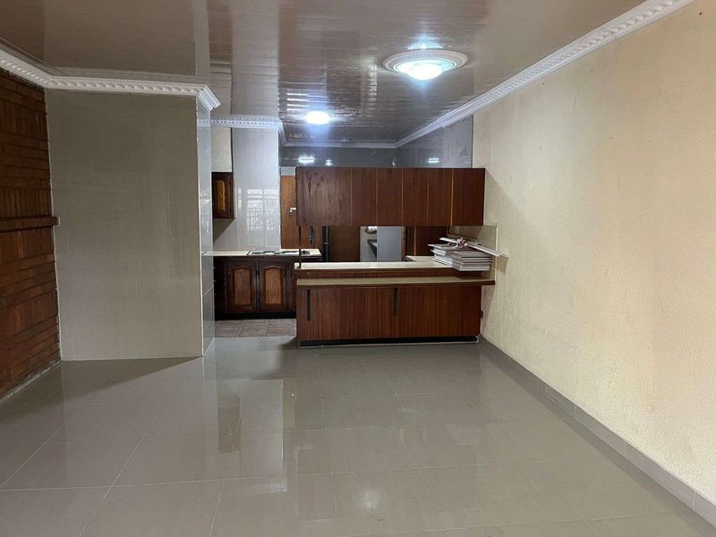 An open-plan living and kitchen area featuring tiled flooring, a breakfast bar, and traditional wooden cabinetry in need of modernization.