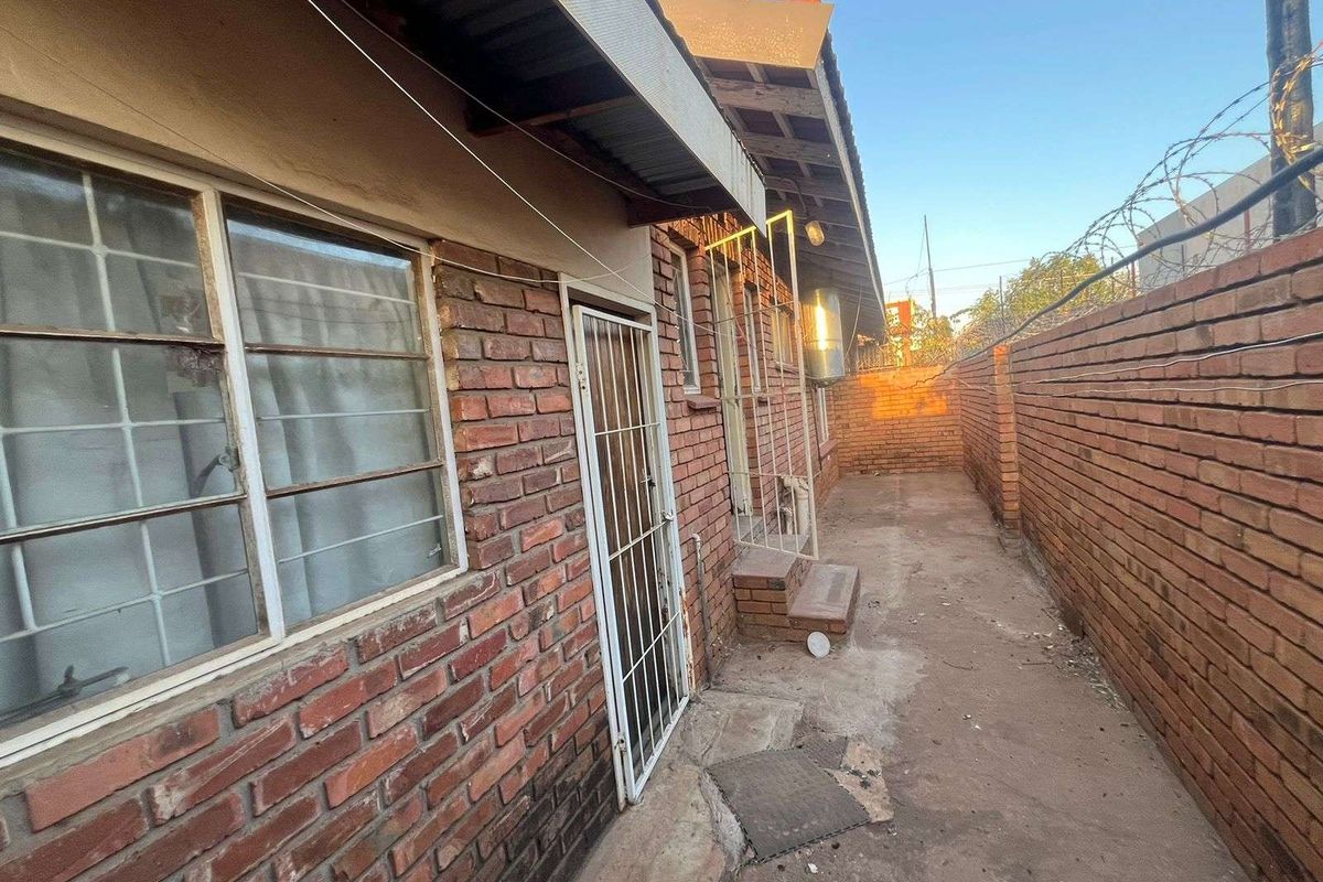 A narrow exterior side passage featuring exposed brick walls, concrete paving, and metal security gates, providing access to the rear of the townhouse.