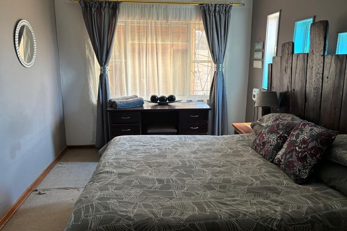 A bedroom featuring a large window with curtains and a custom wooden headboard, showing signs of wear on the flooring.