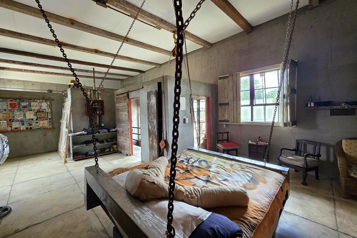 A rustic bedroom featuring exposed concrete walls, timber ceiling beams, and a unique suspended bed frame with industrial chain supports.
