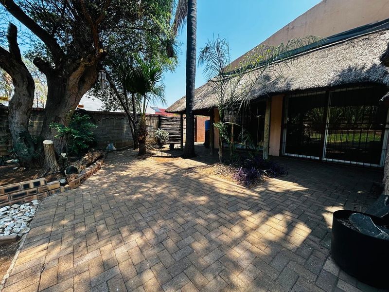 A rear garden area featuring a paved patio, mature trees, and a thatched roof structure with sliding glass door access.