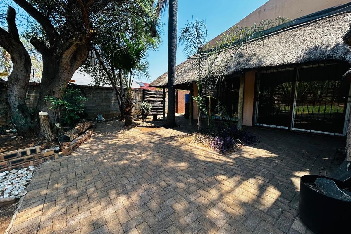 A rear garden area featuring a paved patio, mature trees, and a thatched roof structure with sliding glass door access.