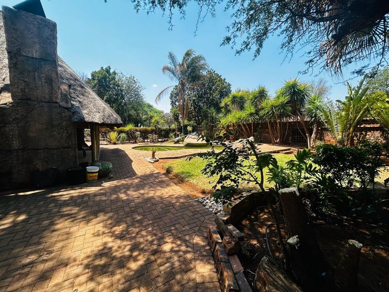 Outdoor pool area featuring a thatched roof structure, stone chimney, and brick paving, set within a mature garden with boundary walls.