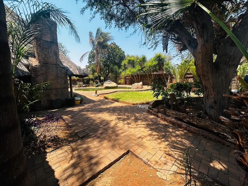 A spacious garden area featuring brick paving, a thatched outdoor structure with a stone chimney, and a swimming pool surrounded by mature trees.