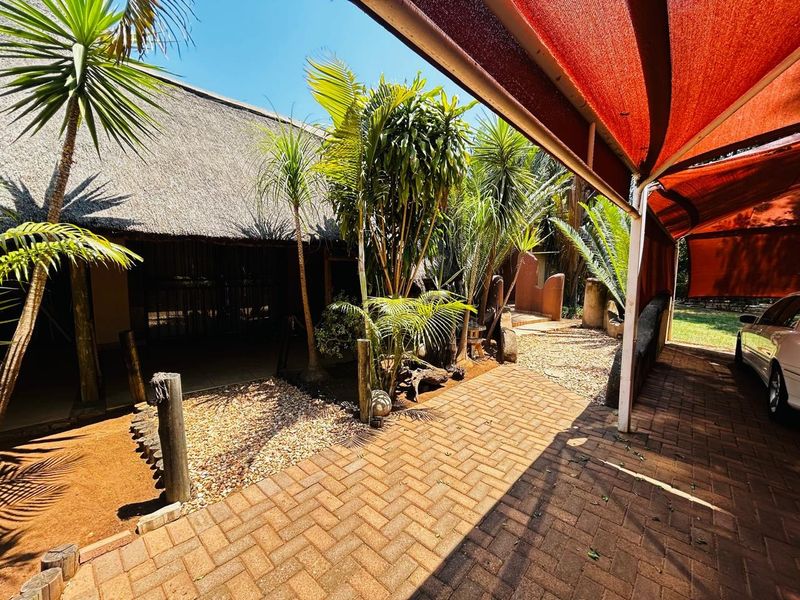 A covered outdoor patio area featuring a thatched roof, paved flooring, and a large red shade sail providing protection for the driveway area.