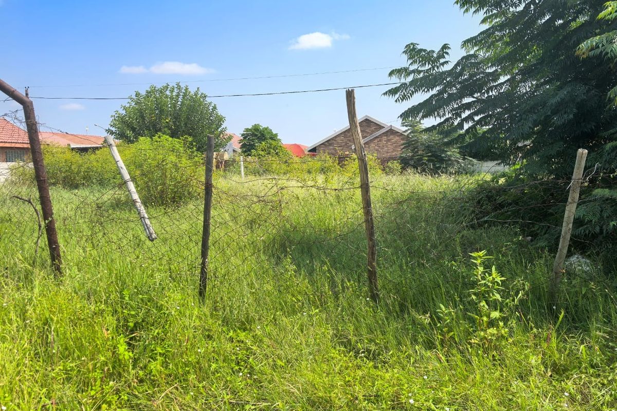 A vacant residential plot featuring overgrown grass and basic wire mesh perimeter fencing with weathered wooden posts.