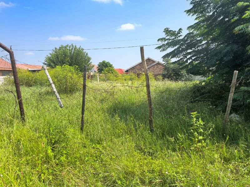 A vacant residential plot featuring overgrown grass and basic wire mesh perimeter fencing with weathered wooden posts.