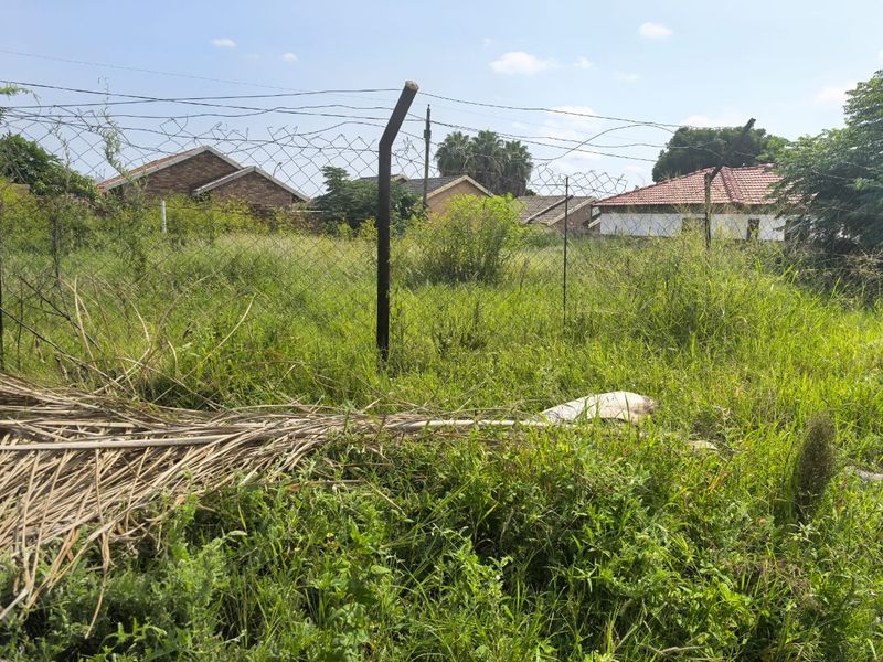 A view of a vacant residential plot featuring overgrown grass and existing wire mesh perimeter fencing.