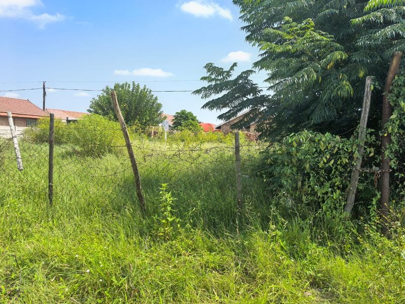 A view of a vacant residential plot featuring overgrown grass, mature trees, and existing wire mesh perimeter fencing.