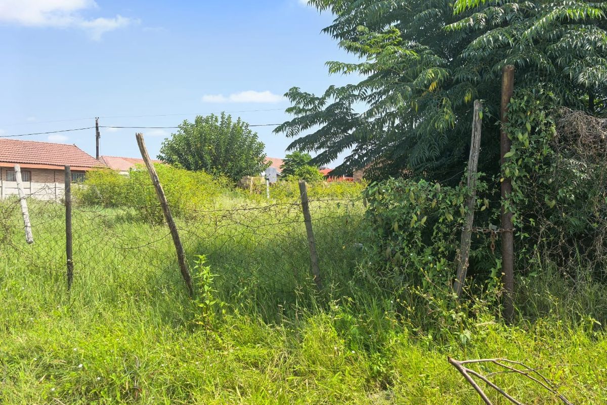 A view of a vacant residential plot featuring overgrown grass and damaged wire mesh perimeter fencing.