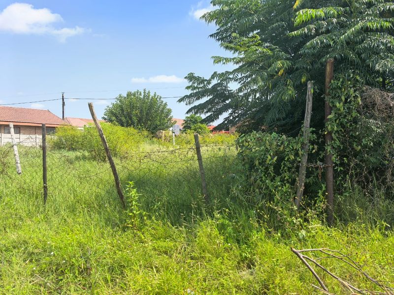 A view of a vacant residential plot featuring overgrown grass and damaged wire mesh perimeter fencing.