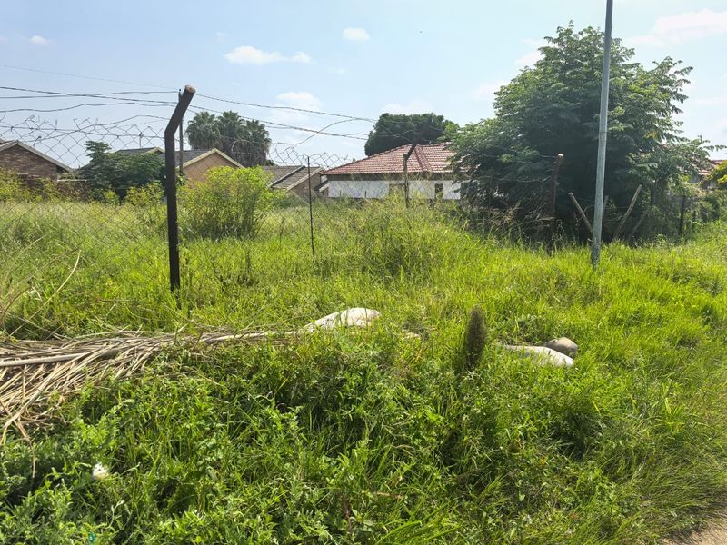 A vacant residential plot featuring overgrown grass and existing wire mesh perimeter fencing with metal posts.