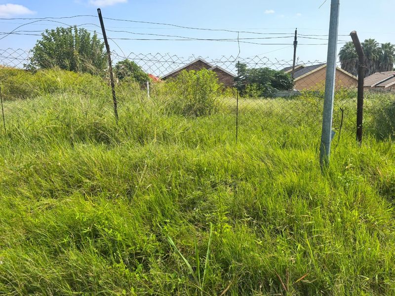 A view of a vacant residential plot featuring overgrown grass and perimeter wire mesh fencing.