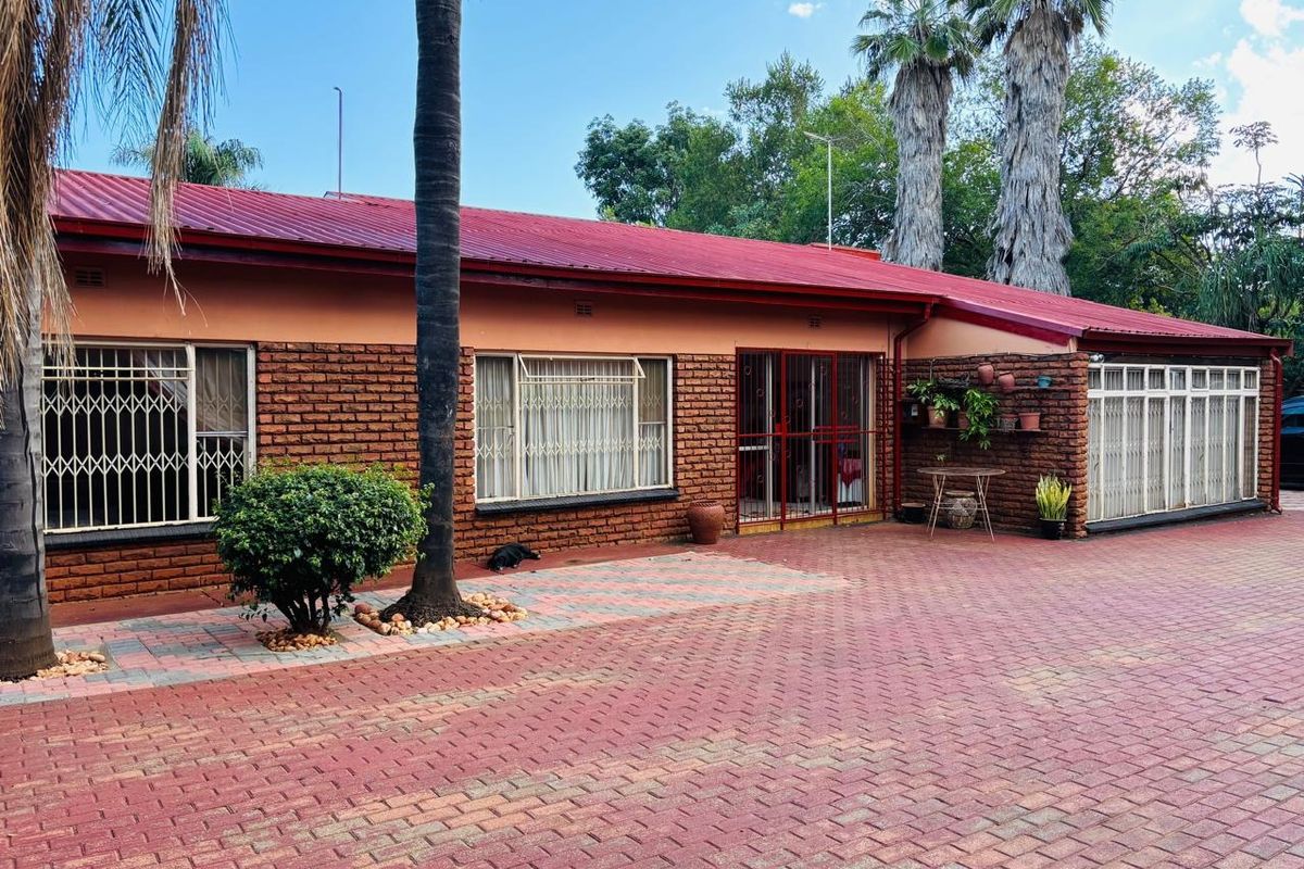 Rear view of a single-story brick house featuring a red metal roof, security gates, and an extensive paved courtyard area.